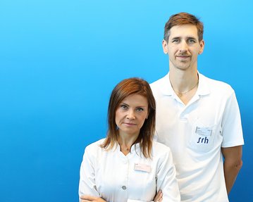 Zwei Fachkräfte in weißen Kitteln stehen vor einem blauen Hintergrund. Die Frau hat lange, braune Haare und der Mann hat kurzes, braunes Haar.