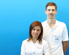 Zwei Fachkräfte in weißen Kitteln stehen vor einem blauen Hintergrund. Die Frau hat lange, braune Haare und der Mann hat kurzes, braunes Haar.