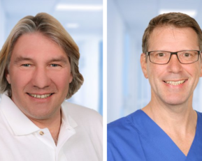 Zwei Männer in professioneller Kleidung, einer in einem weißen Polohemd und der andere in einem blauen Arztkittel, lächeln in die Kamera.