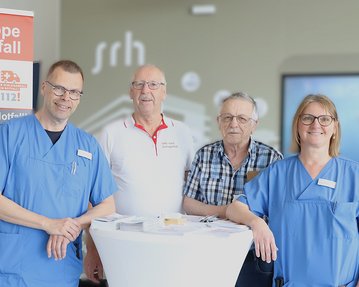 Vier Personen stehen an einem Tisch, umgeben von einem Informationsbanner zur Selbsthilfegruppe nach Schlaganfall. Sie tragen medizinische Kleidung.