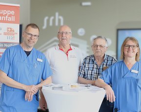 Vier Personen stehen an einem Tisch, umgeben von einem Informationsbanner zur Selbsthilfegruppe nach Schlaganfall. Sie tragen medizinische Kleidung.