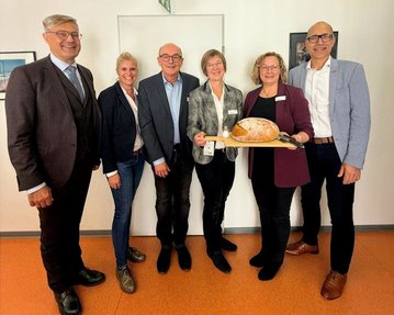 Sechs Personen stehen zusammen und halten ein großes Brot auf einem Tablett. Sie tragen formelle Kleidung und lächeln.