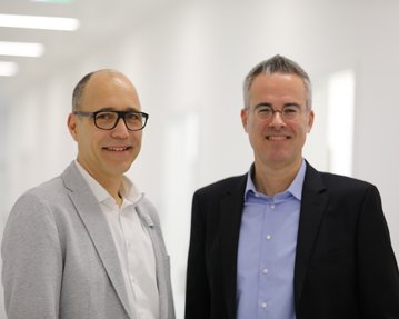 Zwei Männer stehen in einem hellen Flur. Der Mann links trägt eine Brille und einen grauen Blazer, der Mann rechts hat ein schwarzes Sakko und ein blaues Hemd an.
