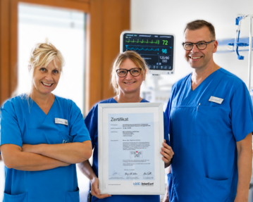 Drei medizinische Fachkräfte in blauen Uniformen halten ein Zertifikat in der Mitte. Im Hintergrund sind medizinische Geräte sichtbar.