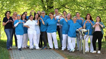 Eine Gruppe von Pflegekräften in blauen Uniformen formt mit ihren Händen Herzen. Sie stehen auf einem Gehweg umgeben von Bäumen.