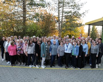 Eine große Gruppe von Menschen steht im Freien, umgeben von Bäumen, und posiert für ein Gruppenfoto. Es sind überwiegend Frauen zu sehen.