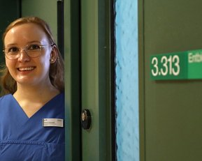 Eine Person in blauer Arbeitskleidung steht an einer Tür mit der Nummer 3.313 und lächelt freundlich.