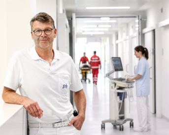 Ein Arzt in weißer Kleidung steht an einer Wand in einem Krankenhausflur, während im Hintergrund medizinisches Personal und Patienten behandelt werden.