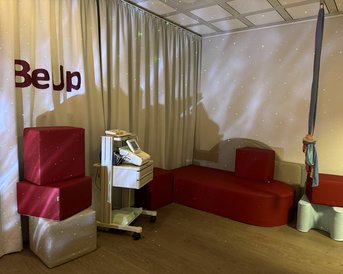 Ein Raum mit roten Sitzmöbeln, einem medizinischen Gerät und einem Vorhang mit der Aufschrift "BeUp".